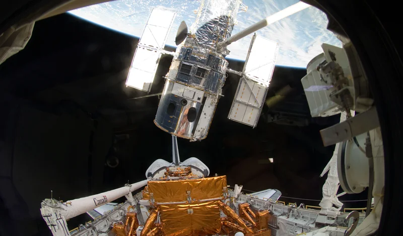 Telescopul spațial Hubble ținut de brațul robotic al navetei, deasupra Pământului