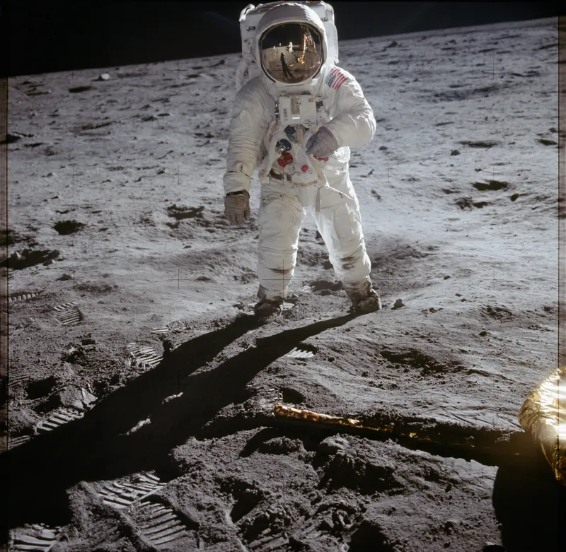 Buzz Aldrin în costum spațial alb, mergând pe solul cenușiu al Lunii lângă modulul lunar, iulie 1969